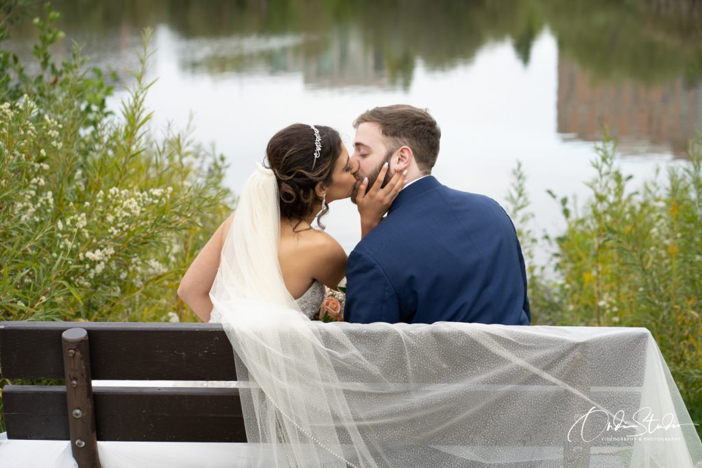 10 Essential Tips for Choosing the Perfect Wedding Photographers in Toronto 74 This romantic photo, captured by ONDI Studio, shows a couple sharing a tender kiss while seated on a bench by a peaceful lakeside. The bride, with her veil elegantly draped over the bench, holds her groom’s face as they embrace in this quiet moment. The serene water and lush greenery in the background create a natural and intimate setting, perfectly complementing the couple’s love and joy. The image captures the essence of romance and the beauty of the surroundings, making it a timeless wedding memory.