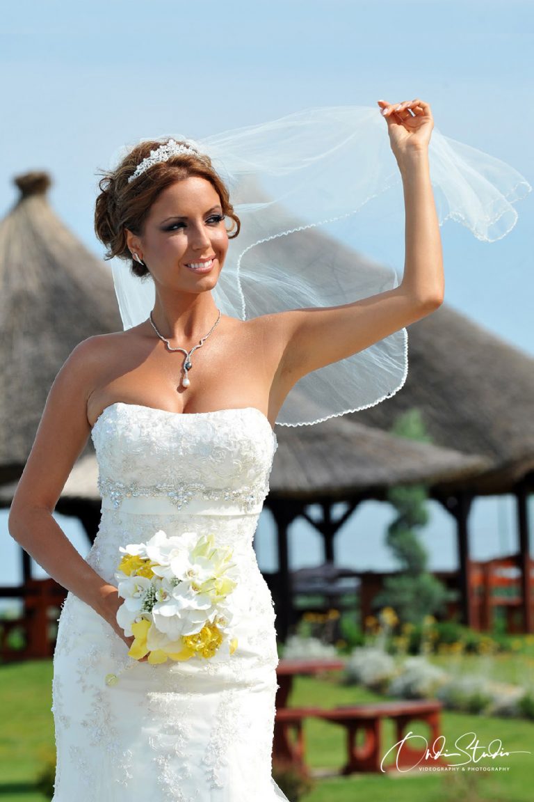 A bride in an elegant strapless wedding gown holds her veil in the breeze, with a bouquet of white and yellow flowers, standing in a sunny garden gazebo.