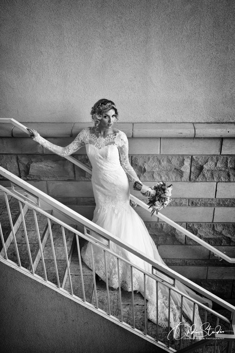 A black and white photograph captures a bride in a lace-detailed gown standing on a staircase, looking pensively into the distance.