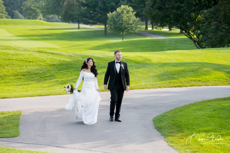 A joyful bride and groom walk hand in hand across a lush golf course, the bride holding her dress and bouquet, with trees and greenery in the background.