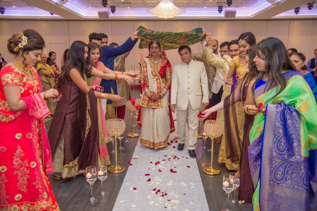 Unforgettable Indian Weddings: Top Photographers and Videographers in Toronto & GTA 6 The image shows a traditional Indian wedding ceremony. The bride, dressed in an ornate red and white bridal lehenga with intricate jewelry, walks down the aisle accompanied by a man, likely a family member. The bride's path is lined with rose petals, and she is shaded by a green cloth held by attendants. Women in colorful sarees and dresses flank the aisle, some scattering petals. The setting is elegant, with candles in glass holders and floral decor, creating a festive and sacred atmosphere. Photo Taken by Ondi Studio
