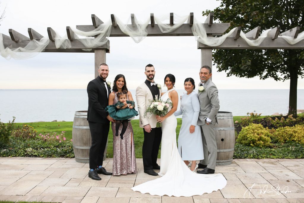 Affordable Wedding Videographer in Toronto & GTA | ONDI STUDIO 15 Valeria and Matt's wedding, beautifully captured by ONDI Studio, showcases an outdoor setting by the waterfront. The couple stands under a wooden pergola draped with delicate fabric, with Valeria in a stunning white gown and Matt in a light-colored tuxedo. They are surrounded by family members, including a young child, all dressed elegantly. The peaceful ocean in the background adds a romantic touch to the moment, perfectly encapsulated by ONDI Studio.