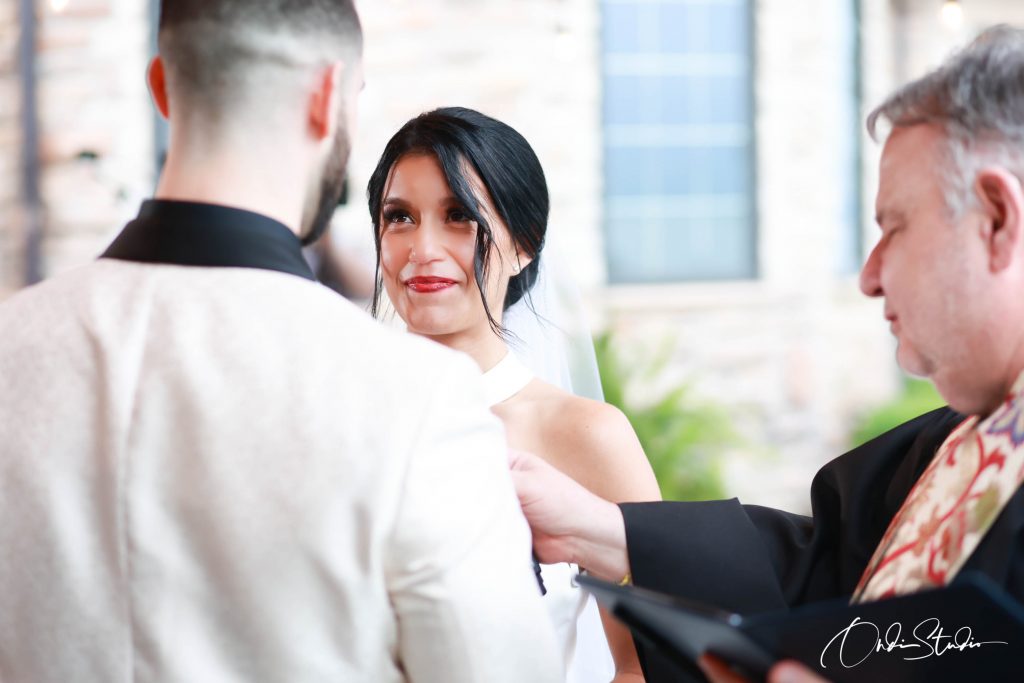 The Ultimate Guide to Finding the Perfect Wedding Photographer in Toronto & GTA 1 Valeria looks lovingly at Matt during their wedding ceremony in an industrial chic venue. The officiant reads from a book in the foreground. Photo by ONDI Studio.