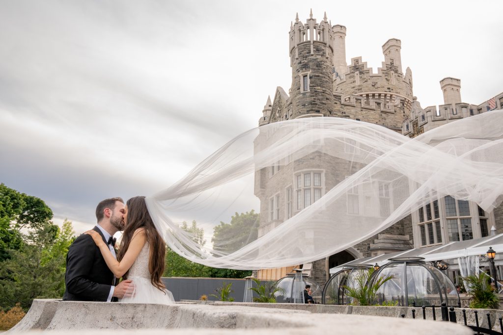 The Impact of Wedding Photography on Social Media 39 Bride and groom share a kiss on a balcony, with the bride's veil elegantly caught in the breeze, framing a majestic castle in the background. Photo captured by ONDI Studio.